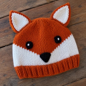 May include: A knitted beanie hat designed to resemble a fox, featuring a vibrant orange base with a white lower section and a ribbed brim. The hat includes two pointed ears with white centers, two round black eyes, and a small black triangular nose.