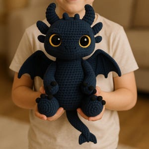 Puede incluir: Un peluche de dragón tejido en color azul oscuro con grandes ojos amarillos y detalles negros. El dragón tiene pequeños cuernos, alas y una larga cola. El juguete está siendo sostenido por una persona que lleva una camiseta clara.