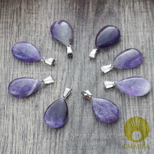 May include: Seven teardrop-shaped amethyst pendants with silver-colored bails are arranged in a circular pattern. The pendants display varying shades of purple and are set against a gray wooden surface. The pendants are likely for jewelry making.