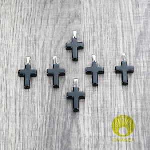 May include: A collection of six black cross pendants with silver-colored bails. The crosses are made of a smooth, dark material and are arranged on a light gray wooden surface. The pendants are a religious symbol.