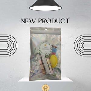 Może przedstawiać: Przezroczysta, opalizująca torebka z napisem "Anti-Panic Pack" zawiera różne przedmioty, w tym brelok, balsam do ust i żółty przedmiot. Nad torebką widnieje napis "NEW PRODUCT". Na dole znajduje się logo Luminosity Learning.