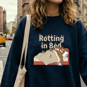 Puede incluir: Sudadera azul marino con la frase "Rotting in Bed" y una ilustración de un perezoso en la cama. El perezoso es marrón y beige, descansando bajo una manta beige en una cama roja. La sudadera la lleva una persona con una gorra beige.