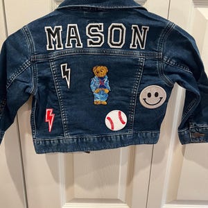 May include: A dark blue denim jacket featuring the name MASON in white block letters across the back. The jacket is decorated with several patches, including a brown teddy bear, a white baseball, a silver glitter smiley face, and two lightning bolts.