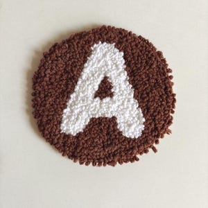 May include: A brown and white round rug with the letter A in white yarn on a white background.