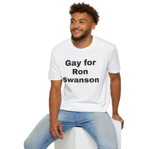May include: A white t-shirt with the text "Gay for Ron Swanson" in black. The shirt is worn by a person in blue jeans. The person is sitting on a white surface.