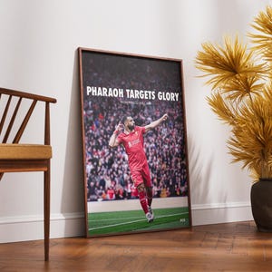 May include: A framed poster featuring a soccer player in a red uniform, with the text "PHARAOH TARGETS GLORY" and the player's name. The image shows the player celebrating on a green field in front of a crowd. The frame is brown.