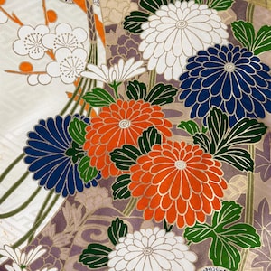 以下が含まれることがあります： オレンジ、白、濃紺の大きな菊の花、緑の葉、白い梅の花が描かれた鮮やかな花柄のテキスタイル生地。デザインには、黒と白の斑点模様のテクスチャー部分や幾何学模様が含まれています。
