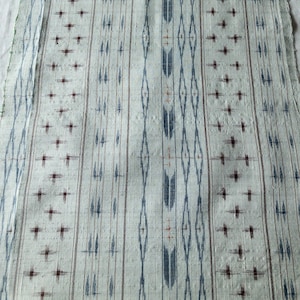May include: A rectangular piece of off-white fabric featuring a vertical pattern of repeating blue and dark red ikat-style motifs. The design consists of alternating stripes of diamond shapes, arrowheads, and small cross-like marks throughout.