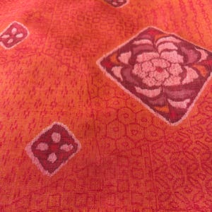 以下が含まれることがあります： 幾何学模様と花柄が繰り返される生地のクローズアップ。生地はオレンジ、赤、ピンクの色合いの鮮やかなカラーパレットが特徴です。デザインにはひし形と花のモチーフが含まれています。