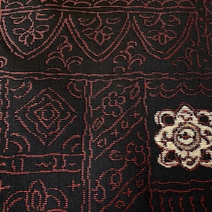 以下が含まれることがあります： 赤色の繰り返し模様の黒いテキスタイルをクローズアップ。デザインには、幾何学的な形、花のモチーフ、様式化された線が含まれています。オフホワイトと茶色の単一の花の要素が見えます。