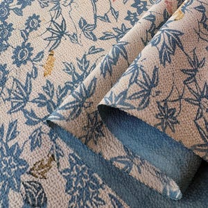 May include: A folded piece of fabric featuring a beige base with a repeating pattern of blue floral and leaf motifs. The textile has a visible woven texture, with small accents of yellow and red scattered throughout the intricate botanical design.