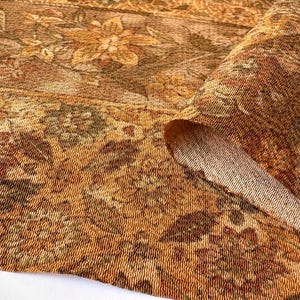 May include: A woven fabric featuring a dense floral pattern in shades of golden yellow, burnt orange, and deep brown. The textile displays a textured surface with intricate botanical motifs and a folded corner revealing the lighter underside.