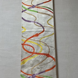 以下が含まれることがあります： 菱形模様のオフホワイトの長方形の織物。オレンジ、黄色、緑、紫、茶色のカラフルなロープのようなデザインが表面に織り込まれています。この織物は装飾品または布地である可能性があります。寸法はセンチメートルです。