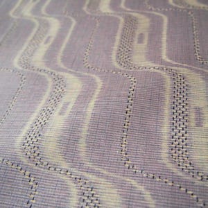 以下が含まれることがあります： ラベンダーとクリーム色の波線模様が繰り返される生地のクローズアップ。デザインには小さな点状のディテールと長方形の形が組み込まれており、テクスチャード加工された外観を作り出しています。生地は微妙な光沢があります。