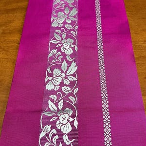 以下が含まれることがあります： マゼンタ色のテーブルランナー。中央に銀色の花と葉の垂直バンドがあり、右側に銀色の幾何学模様があります。テーブルランナーは、銀色の花柄のデザインです。