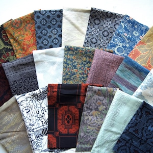 以下が含まれることがあります： さまざまな色と模様の折り畳まれた布の正方形のコレクション。生地には、青、オレンジ、緑、紫の色合いのフローラル、幾何学模様、抽象的なデザインが含まれています。配置は扇状で、さまざまなテキスタイルが展示されています。