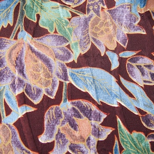 以下が含まれることがあります： 花柄の生地のクローズアップ。デザインは、紫、オレンジ、青の色合いの大きな花を特徴とし、濃いバーガンディ色の背景に配置されています。葉は緑色で、生地は質感のある外観をしています。