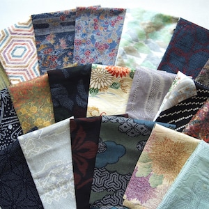 以下が含まれることがあります： さまざまな色と模様の折り畳まれた布の正方形のコレクション。生地は、青、緑、赤、金の色合いで、花柄、幾何学模様、抽象的なデザインを表示します。配置は扇状で、多様性を示しています。