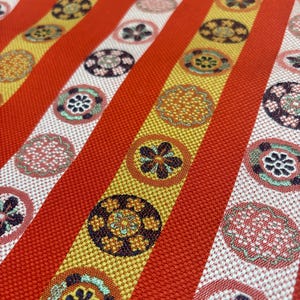 以下が含まれることがあります： 繰り返し模様の生地のクローズアップ。生地は、赤、黄、白のストライプが交互に配置されています。ストライプは、茶色、緑、ピンクなど、さまざまな色の円形のフローラルパターンで飾られています。