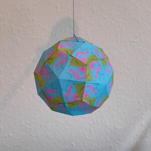 May include: A geometric paper ornament, suspended by a thin black string. The sphere is composed of blue, pink, and green patterned triangular facets. The ornament is hanging against a white wall.