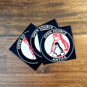 May include: Several black square stickers with a white circular design. The design includes a red penguin and a red Tux the Linux mascot, with the words "OPEN SOURCE" and "ANTIFA" in white.