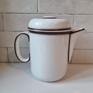 Puede incluir: Tetera de esmalte blanco con tapa y asa curva. La tetera tiene un borde marrón oscuro alrededor de los bordes y el asa. El pico está en el lado derecho. La tetera está sobre una superficie blanca.