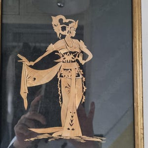 Può includere: Un'opera d'arte incorniciata raffigurante una silhouette dettagliata di una ballerina balinese tradizionale. La figura è realizzata con pezzi di legno chiaro su uno sfondo scuro. La cornice è di un caldo colore dorato.