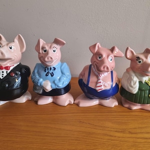 Puede incluir: Un conjunto de cuatro figuras de cerdo de cerámica vestidas con diversos atuendos. El primero lleva un esmoquin negro con pajarita roja. El segundo lleva una blusa azul y falda oscura. El tercero lleva peto azul y corbata a rayas. El cuarto lleva camisa blanca y falda verde.