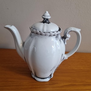 May include: A white ceramic teapot featuring a ribbed body and a matching lid with a finial. The teapot is decorated with dark gray, marble-like smudged accents along the spout, handle, lid rim, and base, creating a distinct contrast against the white.