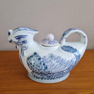Op de afbeelding: Een witte keramische theepot in de vorm van een haan, met blauwe en groene geschilderde details. De theepot heeft een gebogen handvat en een klein deksel met een roze knop. Het ontwerp bevat veer- en schubpatronen.