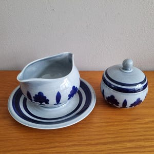 May include: A light blue ceramic gravy boat, saucer, and lidded sugar bowl. The set has a blue tree and leaf design and blue stripes. The items are displayed on a wooden surface.