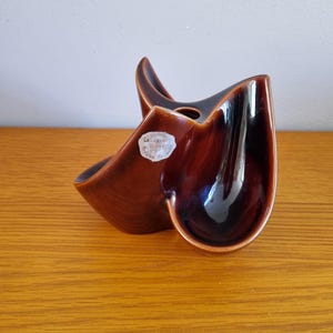 Könnte beinhalten: Eine braune Keramikvase mit glänzendem Finish. Die Vase hat eine einzigartige, asymmetrische Form mit geschwungenen Kanten und einer zentralen Öffnung. Ein kleiner, weißer Aufkleber mit Text ist an der Seite angebracht. Die Vase steht auf einer Holzoberfläche.