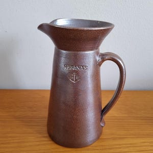 Könnte beinhalten: Ein brauner Keramikkrug mit Henkel und Ausguss. Das Wort "HOGANAS" ist auf der Vorderseite zusammen mit einem Wappen eingeprägt. Der Krug hat eine strukturierte Oberfläche und eine weite Öffnung.