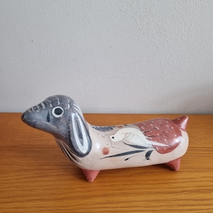 Peut inclure: Une figurine en céramique peinte à la main représentant un chien au corps allongé avec une tête gris foncé, des marques oculaires blanches et un corps beige. Le dos présente un oiseau blanc et des motifs floraux rouges sur des pieds rouges.