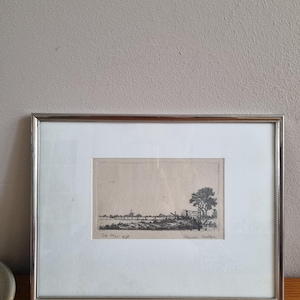 Peut inclure: Une gravure de paysage en noir et blanc encadrée. L'œuvre représente une scène rurale avec un arbre, une clôture et des structures lointaines. Le cadre argenté entoure l'œuvre, qui est placée sur un passe-partout blanc. L'œuvre est signée et datée.