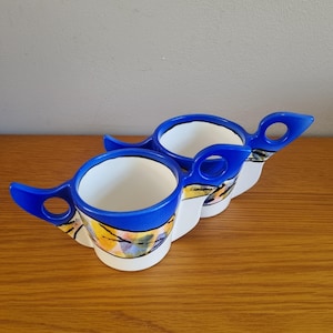 May include: Two ceramic coffee cups with a unique design. The cups have a white interior, blue rims, and blue handles with a hole. The exterior features a colorful abstract pattern with yellow, blue, and black accents. The cups are placed on a wooden surface.