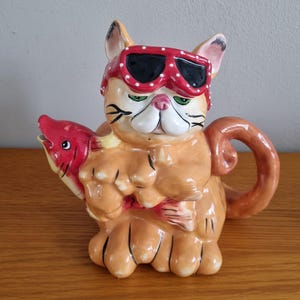 May include: A ceramic cat-shaped teapot in shades of orange and cream, wearing red sunglasses with white polka dots. The cat holds a red fish. The teapot has a brown handle and spout.