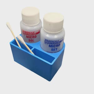 May include: A blue plastic organizer holds two small bottles with white caps and labels that read "MICRO SOL" and "MICRO SET." Two cotton swabs rest in a slot. The bottles are clear, and the labels have red and blue text.