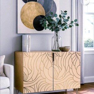 May include: A light wood cabinet with a beige finish and gold leaf design. The cabinet has two doors and sits on thin, gold-colored legs. A painting with gold, black, and gray circles hangs above the cabinet. A glass vase with greenery sits on top.
