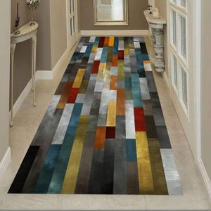 Könnte beinhalten: Ein rechteckiger Flurläufer mit einem geometrischen Muster aus vertikalen Rechtecken in verschiedenen Farben. Der Teppich weist Grautöne, Blau, Gelb, Orange und Rot auf. Der Teppich befindet sich in einem Flur mit weißen Wänden und Türen.