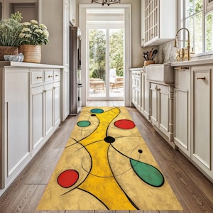May include: A yellow and beige abstract runner rug with red, teal, and black circular and teardrop shapes. The rug is in a kitchen with white cabinets and a wooden floor.