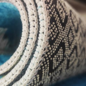 May include: Close-up of a rolled-up rug with a white, textured border. The rug features a geometric pattern in black and white, with a vibrant blue and turquoise interior. The rug's design suggests a focus on home decor and textiles.