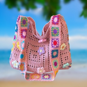May include: A pink crocheted tote bag with a floral design. The bag features a square pattern with colorful floral accents and a strap adorned with more floral squares. The bag is decorated with several 3D flowers.