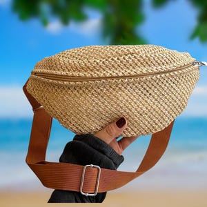 May include: A woven straw fanny pack with a brown zip and adjustable strap. The bag is held up against a backdrop of a beach and ocean. The strap has a silver-coloured buckle.