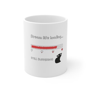 May include: White ceramic mug with the text "Dream life loading..." and "STILL BUFFERING". The design includes a loading bar with hearts and a black cat illustration.