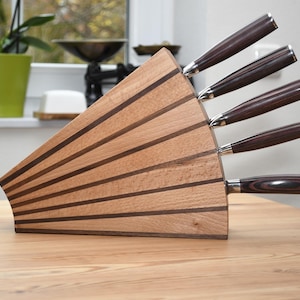 Op de afbeelding: Een houten messenblok met een waaiervormig ontwerp, met afwisselende lichte en donkere houten strepen. Zes messen met donkerbruine handvatten en zilveren accenten zijn in het blok geplaatst.
