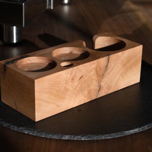 Op de afbeelding: Een lichtgekleurde houten accessoirehouder voor espressomachines met drie cirkelvormige uitsparingen en een kleine ovale uitsparing. De houder staat op een donkere, ronde ondergrond. De houtnerf is zichtbaar.
