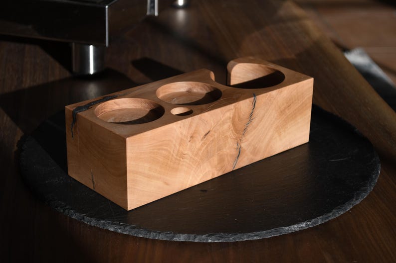 Op de afbeelding: Een lichtgekleurde houten accessoirehouder voor espressomachines met drie cirkelvormige uitsparingen en een kleine ovale uitsparing. De houder staat op een donkere, ronde ondergrond. De houtnerf is zichtbaar.