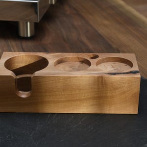 Op de afbeelding: Een houten accessoire voor een espressomachine met een lichtbruine afwerking. Het heeft drie cirkelvormige uitsparingen en een filterhouder. Het accessoire is rechthoekig en staat op een zwart oppervlak.