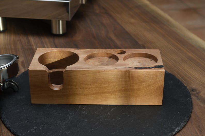 Op de afbeelding: Een houten accessoire voor een espressomachine met een lichtbruine afwerking. Het heeft drie cirkelvormige uitsparingen en een filterhouder. Het accessoire is rechthoekig en staat op een zwart oppervlak.